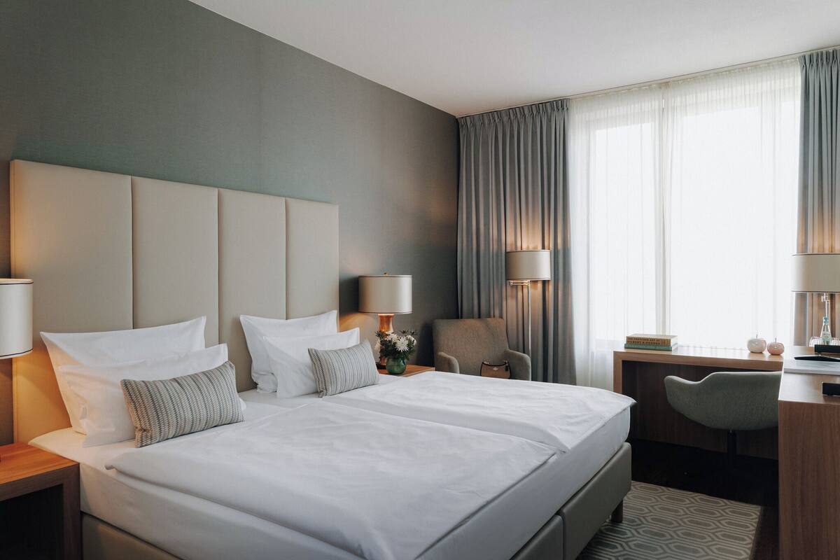 A hotel room with a large bed, two bedside tables, a desk, a chair, and a window with curtains.