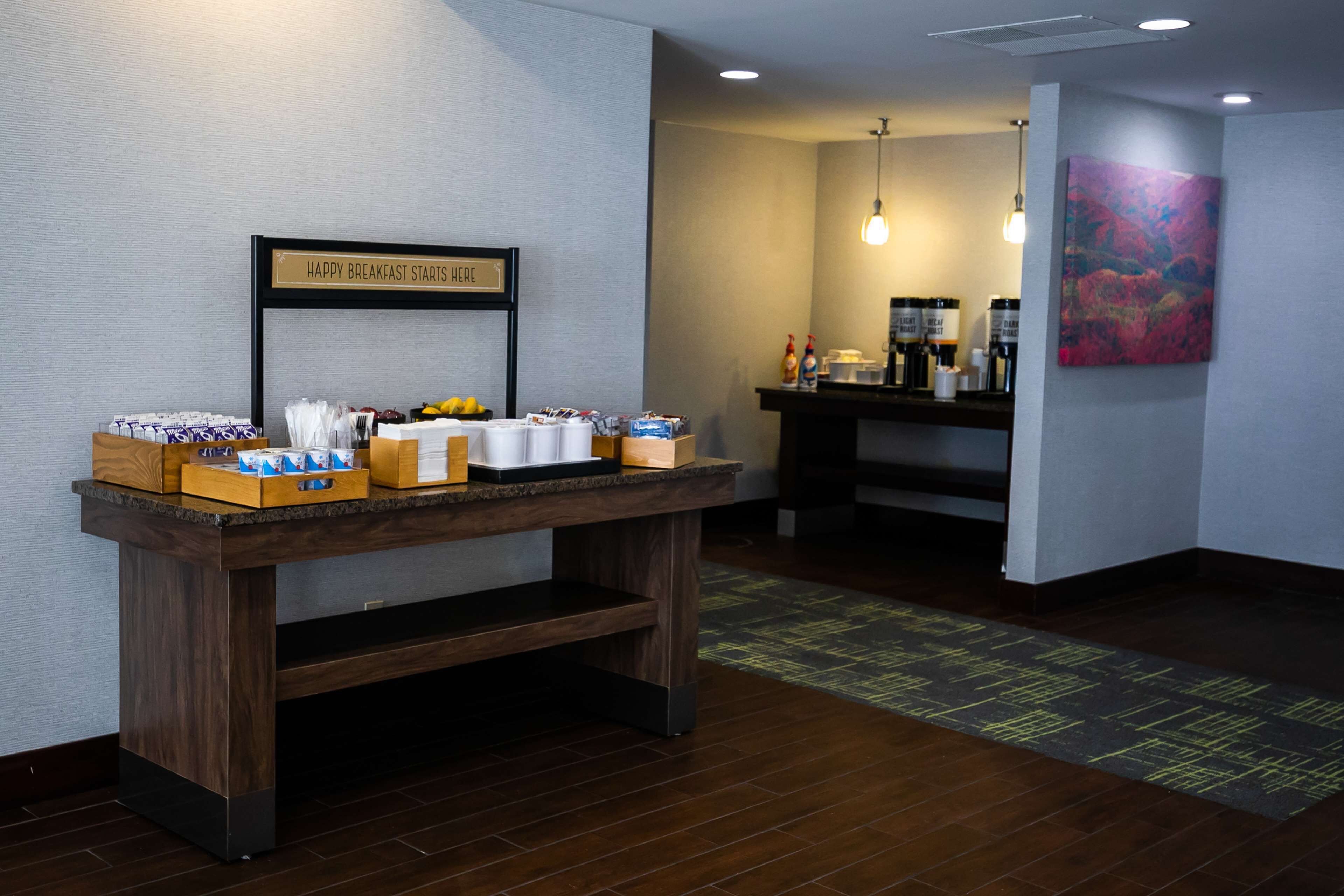 Un buffet de petit-déjeuner avec divers aliments et boissons, un panneau indiquant que le petit-déjeuner commence ici, et un coin café en arrière-plan.