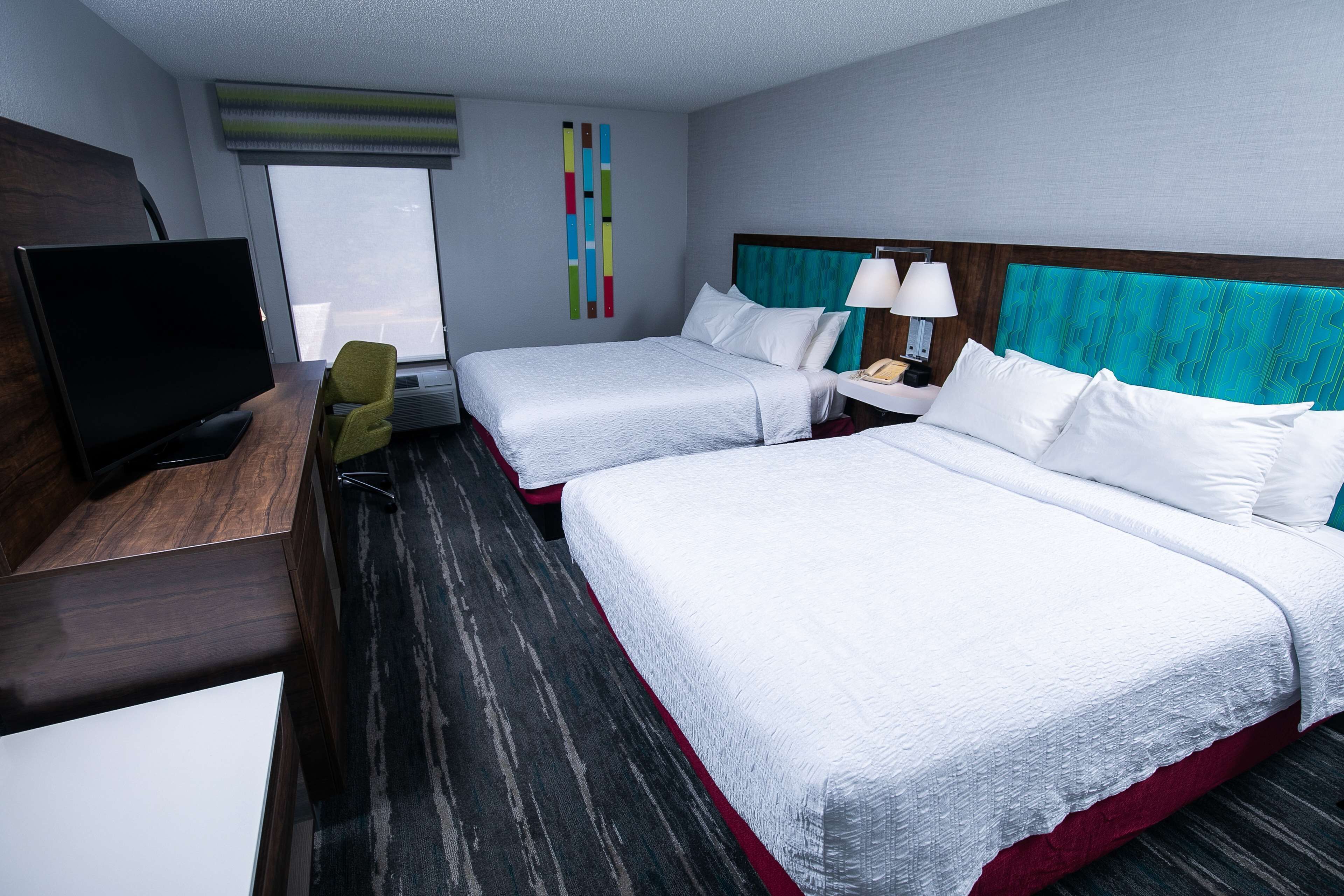 A hotel room with two beds, a TV, a desk, and a window with blinds.
