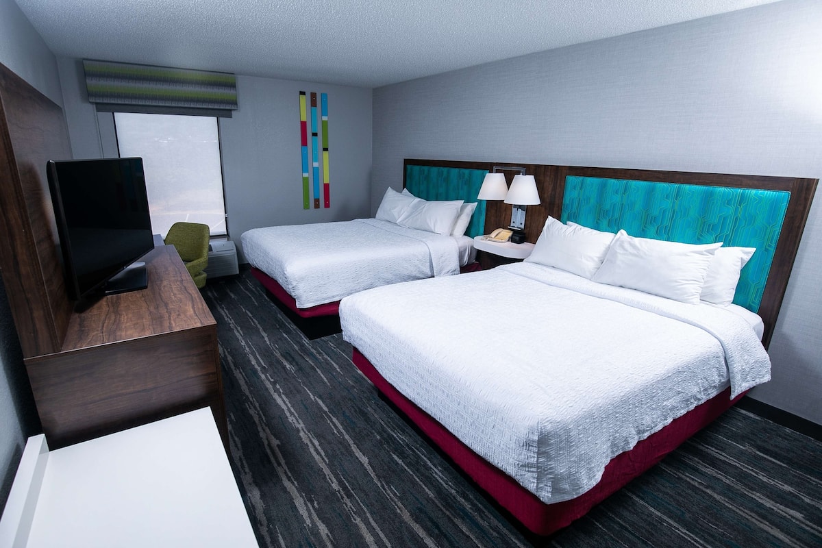 A hotel room with two beds, a TV, a desk, and a window.