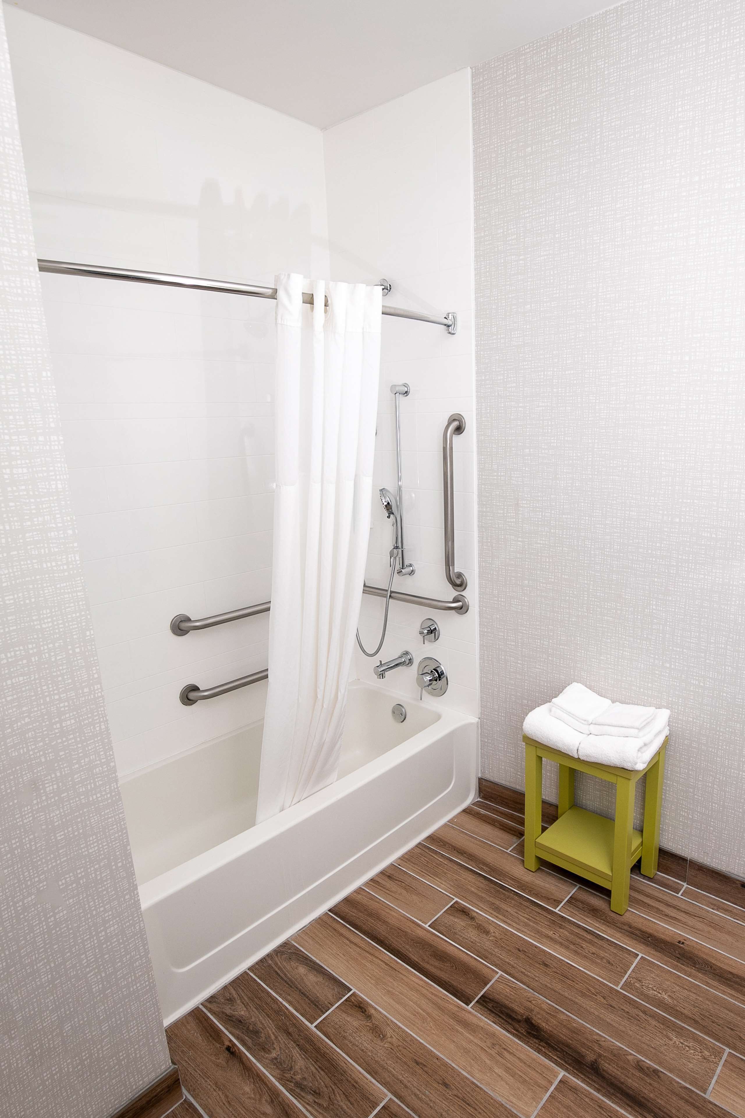 A bathroom with a bathtub, shower, and grab bars. There is a green stool with a towel.