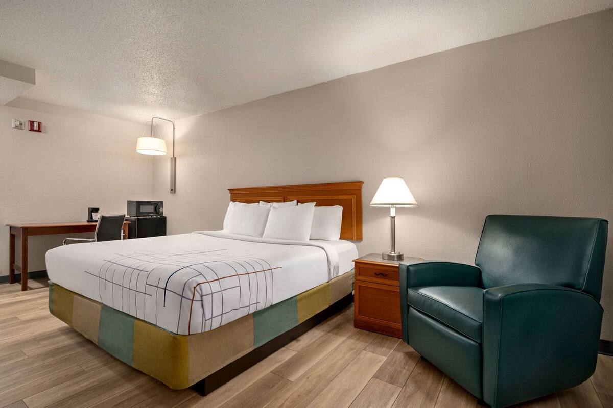 A hotel room with a bed, a desk with a chair, a lamp, and a leather armchair.