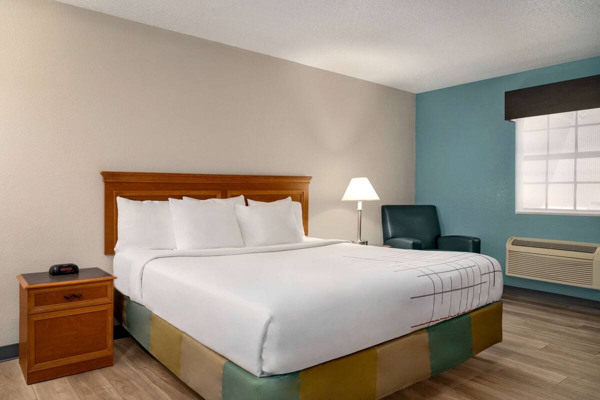 A hotel room with a bed, a nightstand, a desk lamp, a chair, and a window with blinds.