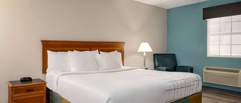 A hotel room with a bed, a nightstand, a desk lamp, a chair, and a window with blinds.