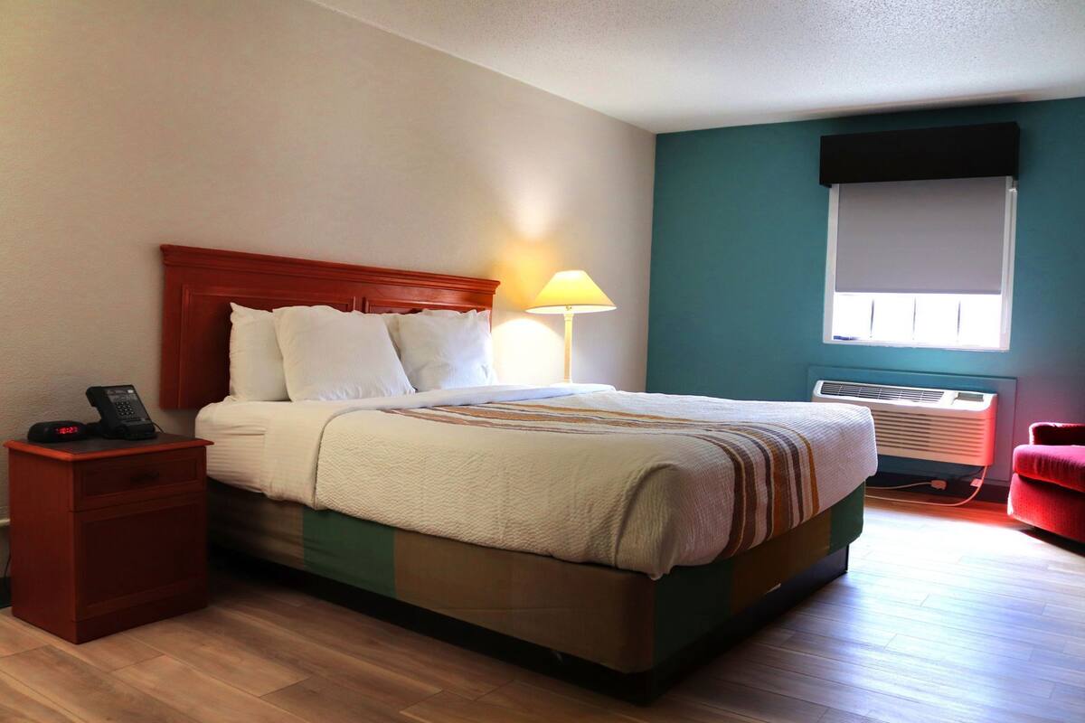 A hotel room with a bed, bedside table, lamp, air conditioning unit, and a window.
