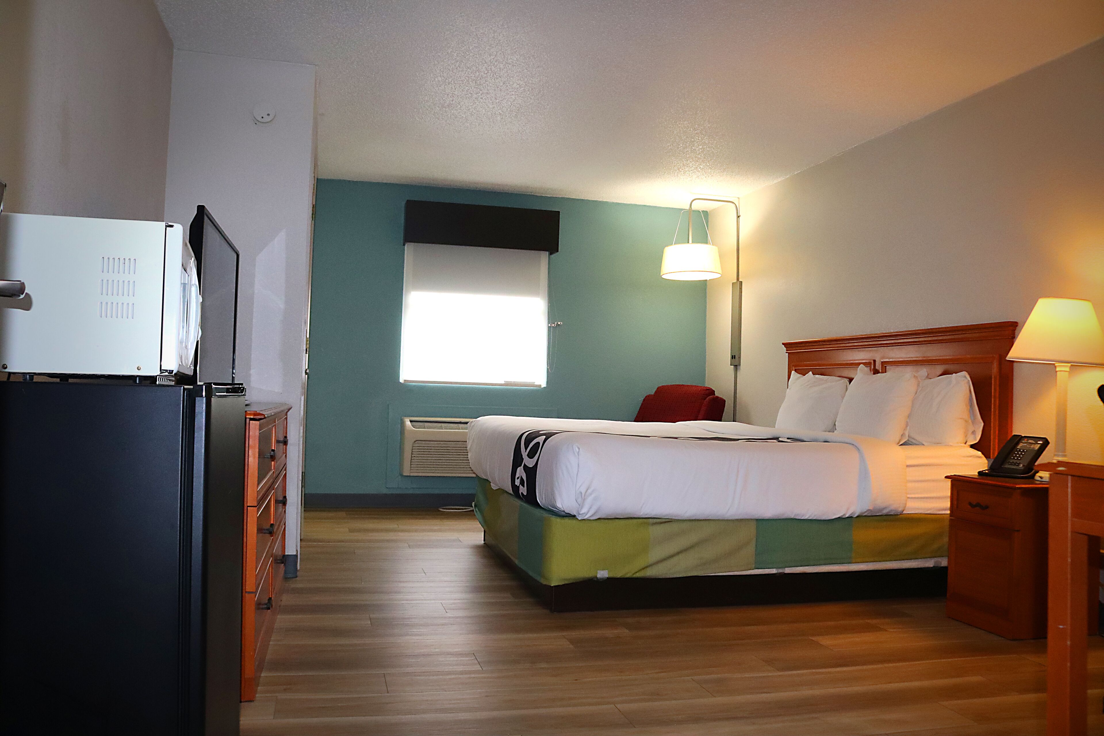 Een hotelkamer met een bed, nachtkastje met lamp, koelkast en raam met jaloezieën.