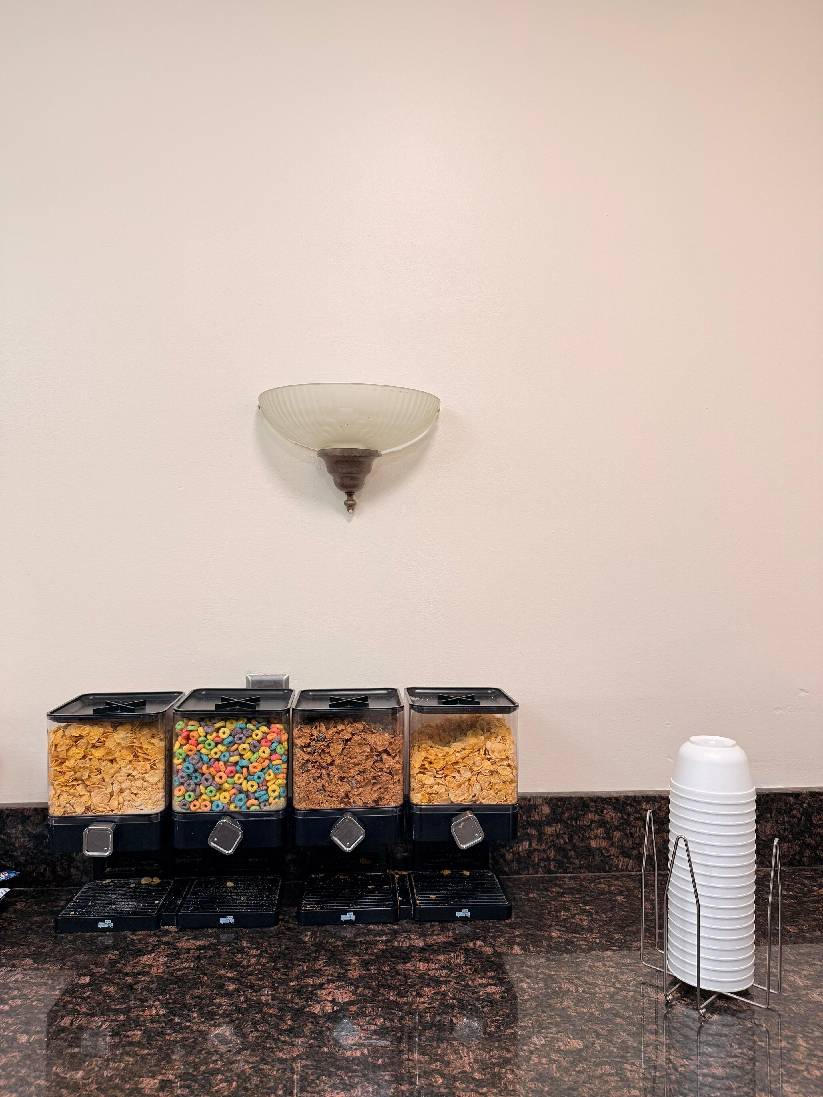 Un mostrador de aperitivos con cuatro recipientes de cereales diferentes y una pila de vasos blancos.