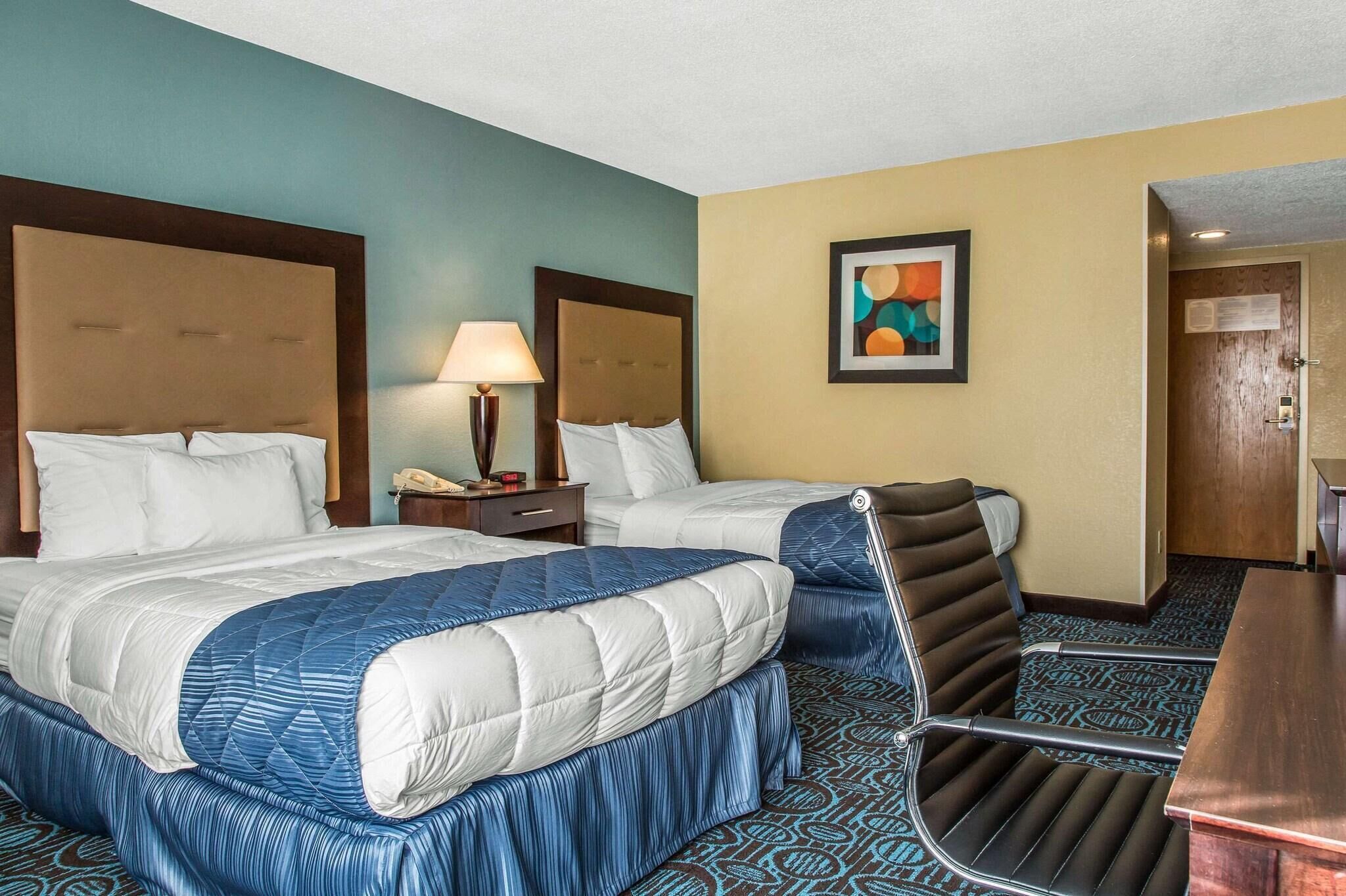 Une chambre d’hôtel avec deux lits, un bureau, une chaise, une lampe et un tableau au mur.