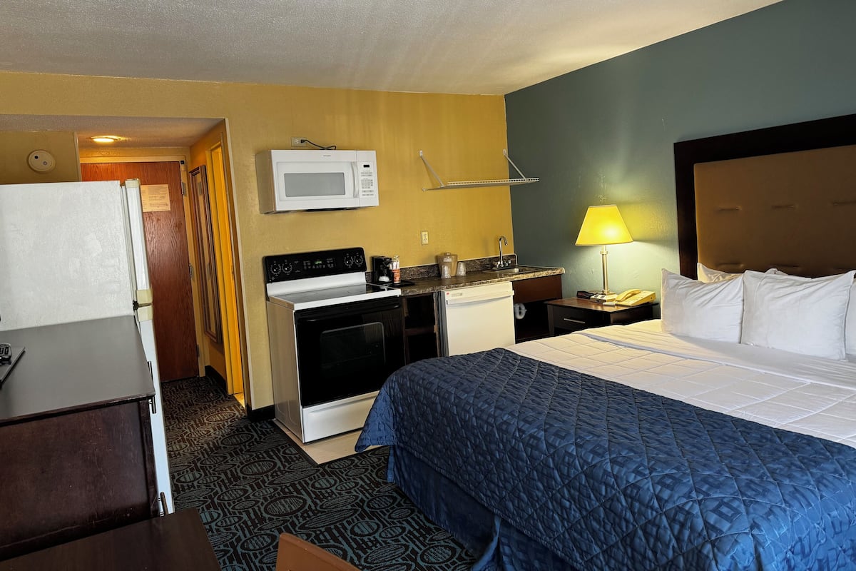 A hotel room with a bed, a microwave, a stove, a refrigerator, a sink, and a nightstand with a lamp.
