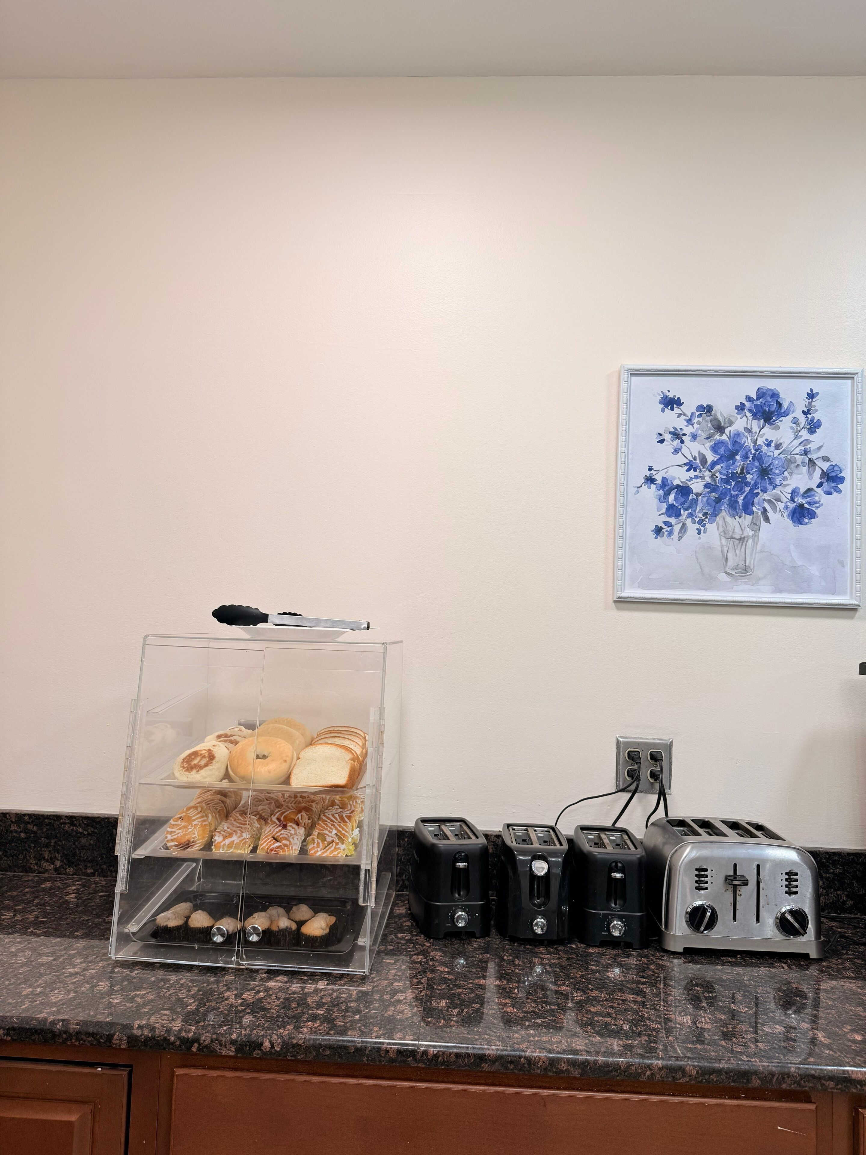 A kitchen counter with a clear display case containing pastries and a toaster. There is a framed painting of flowers on the wall above the counter.