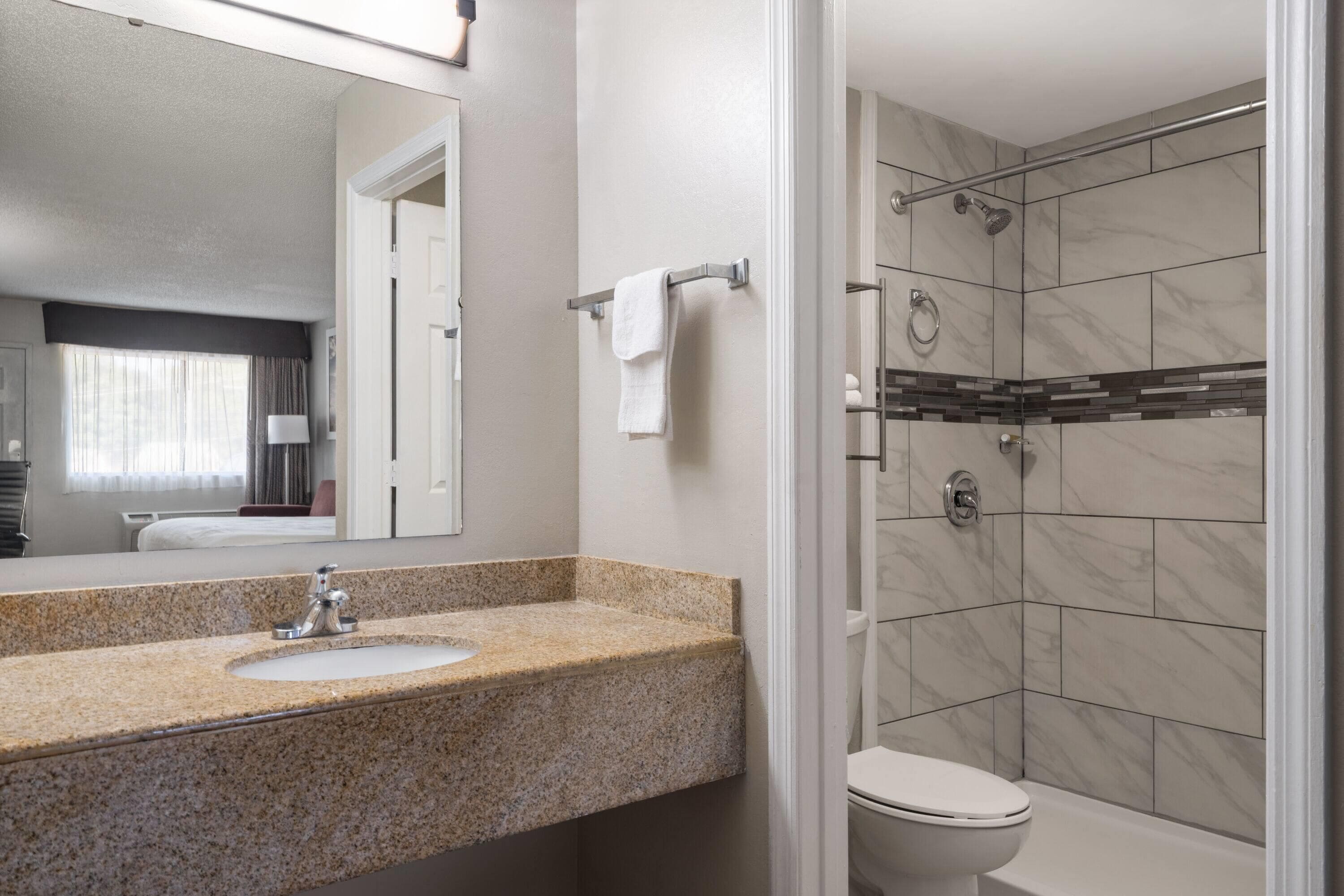 A bathroom with a granite countertop sink, a mirror, a towel rack, a toilet, and a shower with tiled walls.