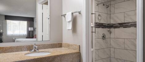 A bathroom with a granite countertop sink, a mirror, a towel rack, a toilet, and a shower with tiled walls.