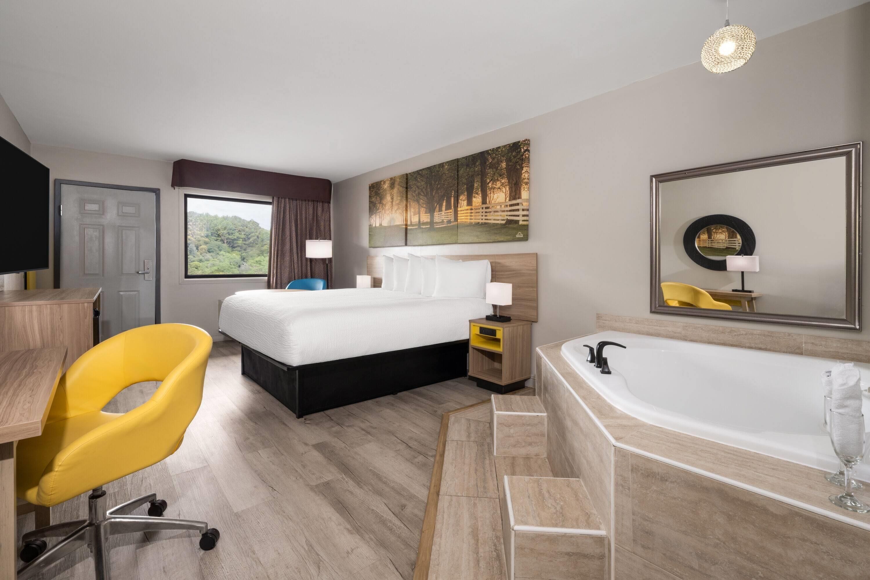 Habitación de hotel con cama, escritorio, silla, baño con bañera y ventana grande con vista a árboles.