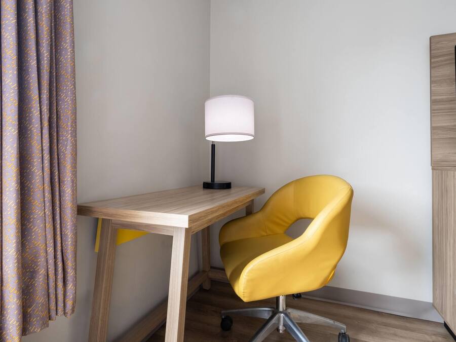 A corner room with a wooden desk, a yellow chair, a white lamp, and a TV mounted on the wall.