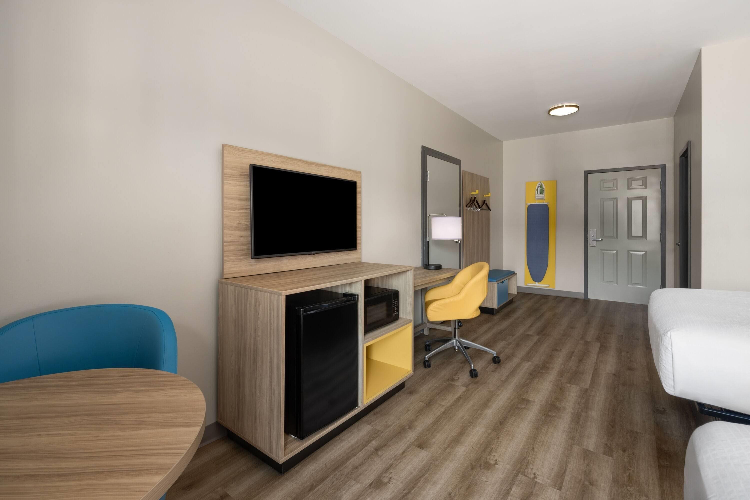 Een hotelkamer met een houten vloer, een aan een houten kast bevestigde televisie, een gele bureaustoel, een wit bed en een deur met een spiegel en kunstwerk.