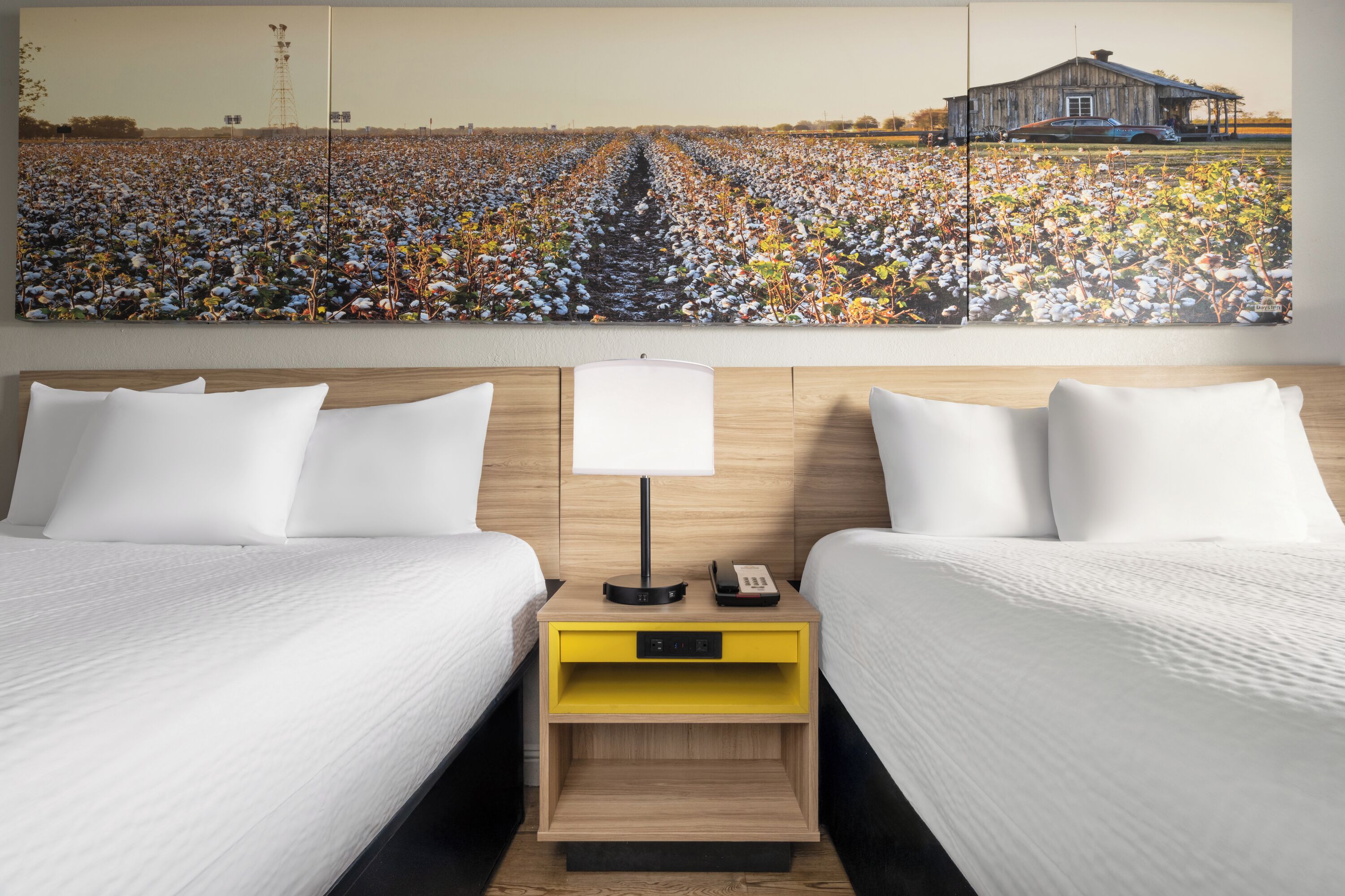 A hotel room with two beds, a nightstand with a lamp, and a large painting of a cotton field.