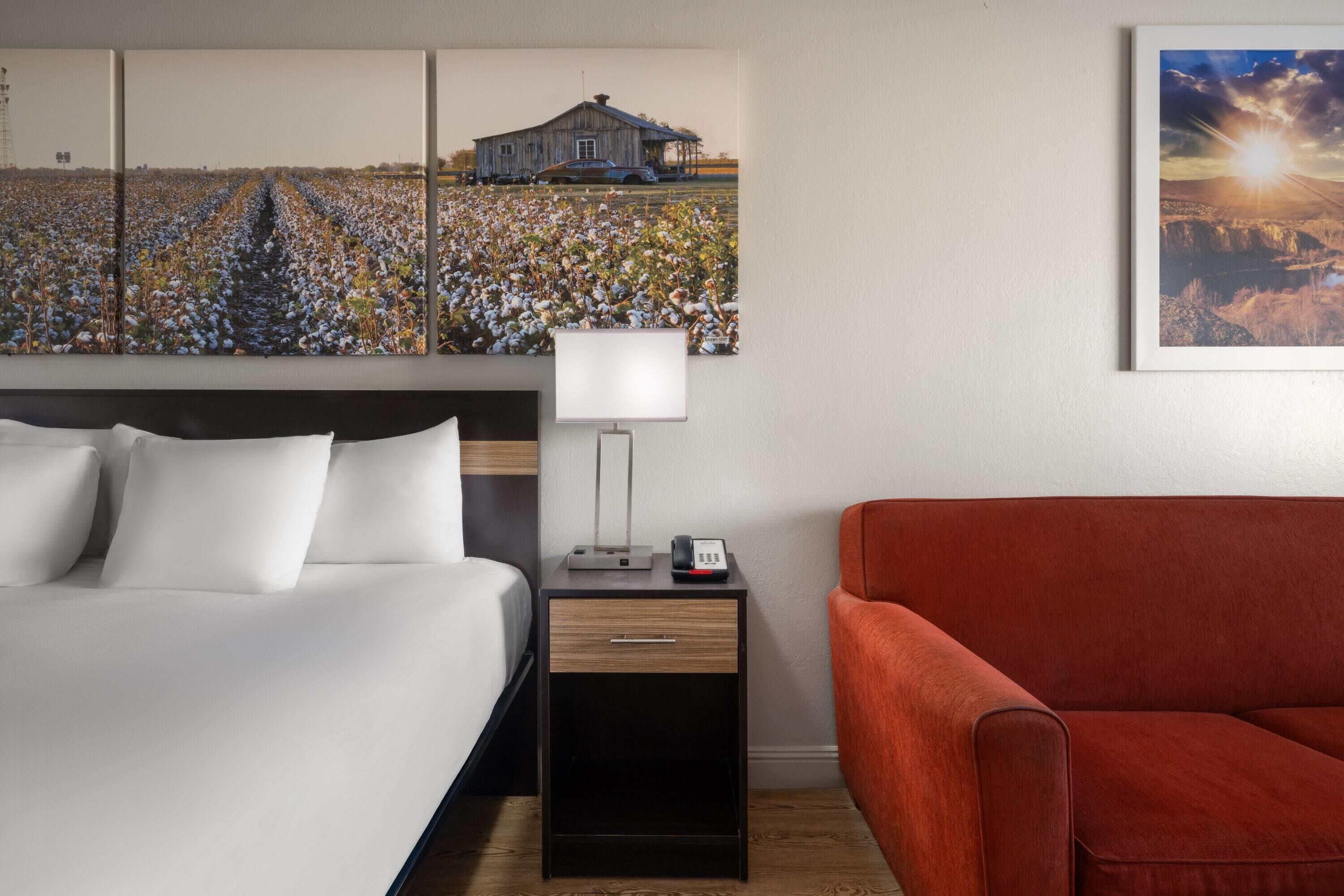 A hotel room with a bed, a nightstand with a lamp, a red sofa, and a large landscape painting on the wall.