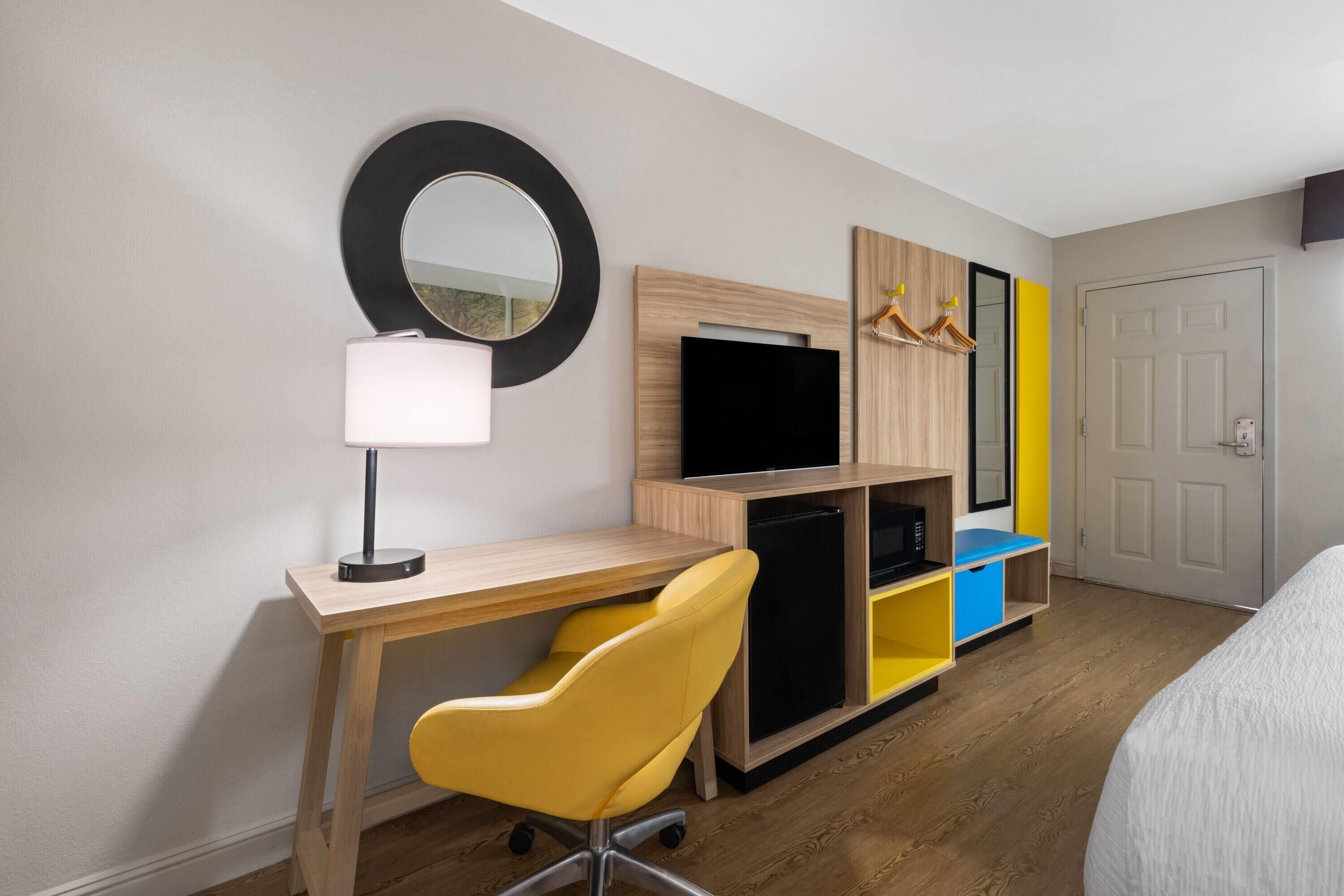 A hotel room with a wooden desk, a yellow chair, a TV, a mirror, and a wardrobe with a mirror and a door.