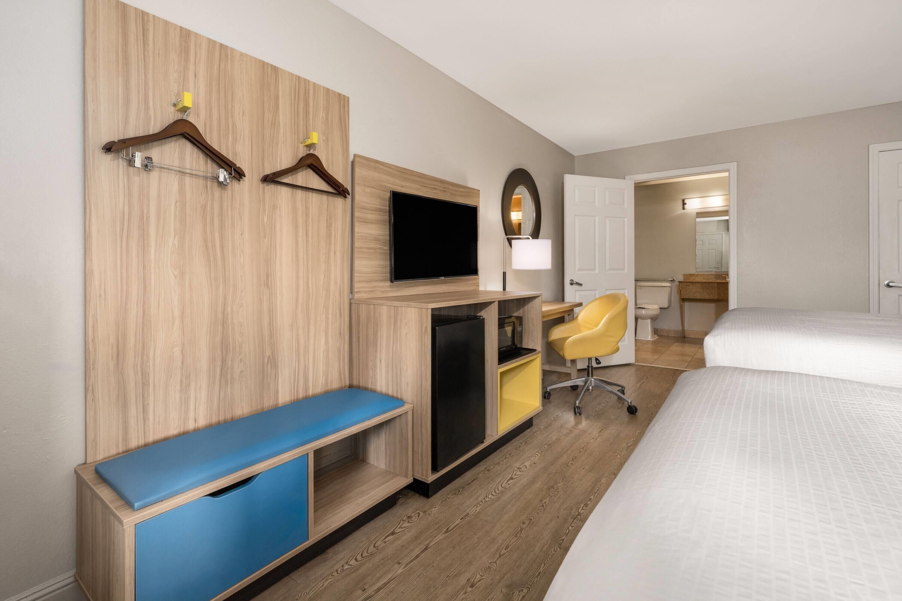 A hotel room with a bed, a desk with a computer, a TV, a mirror, and a bathroom visible through an open door.