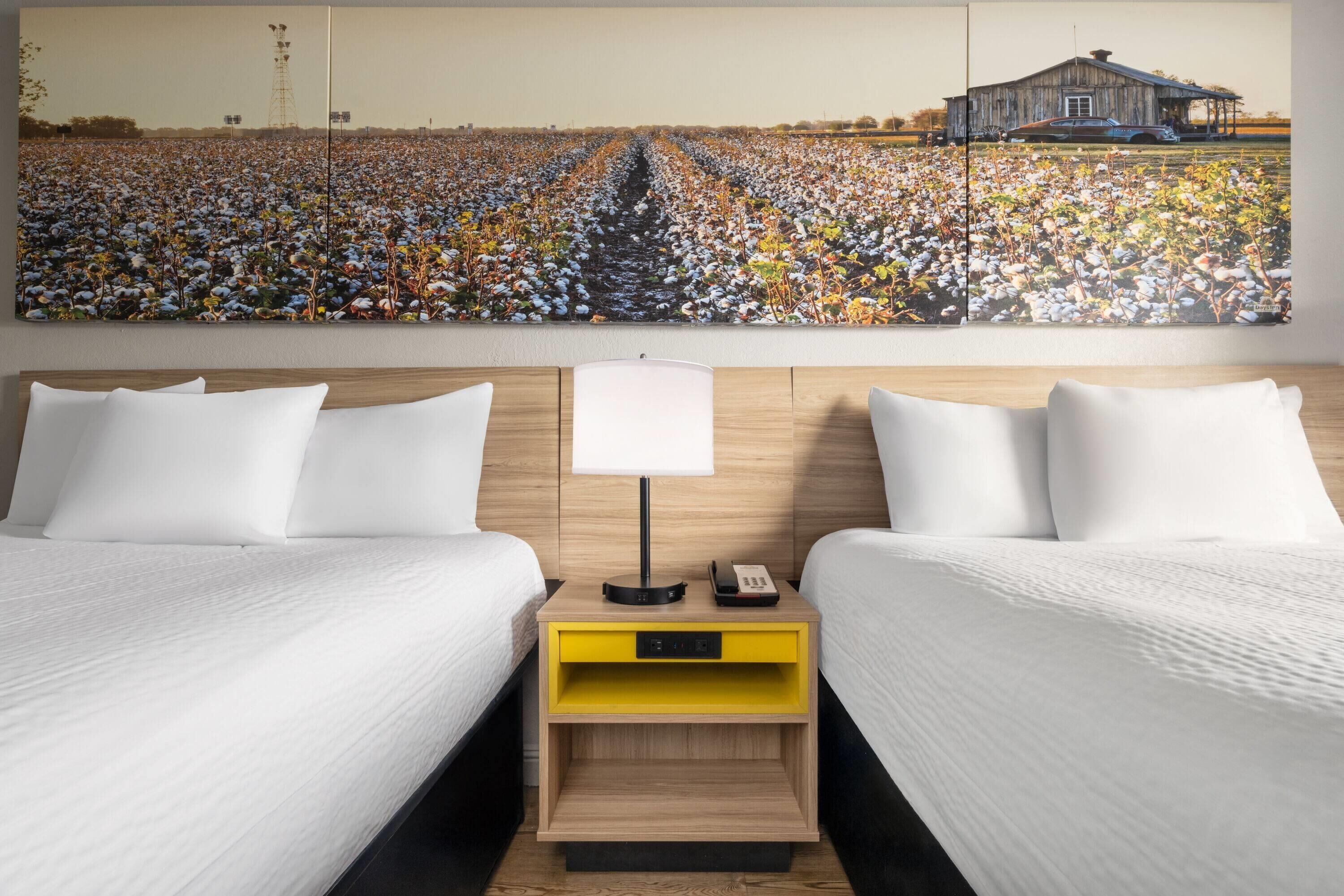 A hotel room with two beds, a nightstand with a lamp, and a large painting of a cotton field and barn.