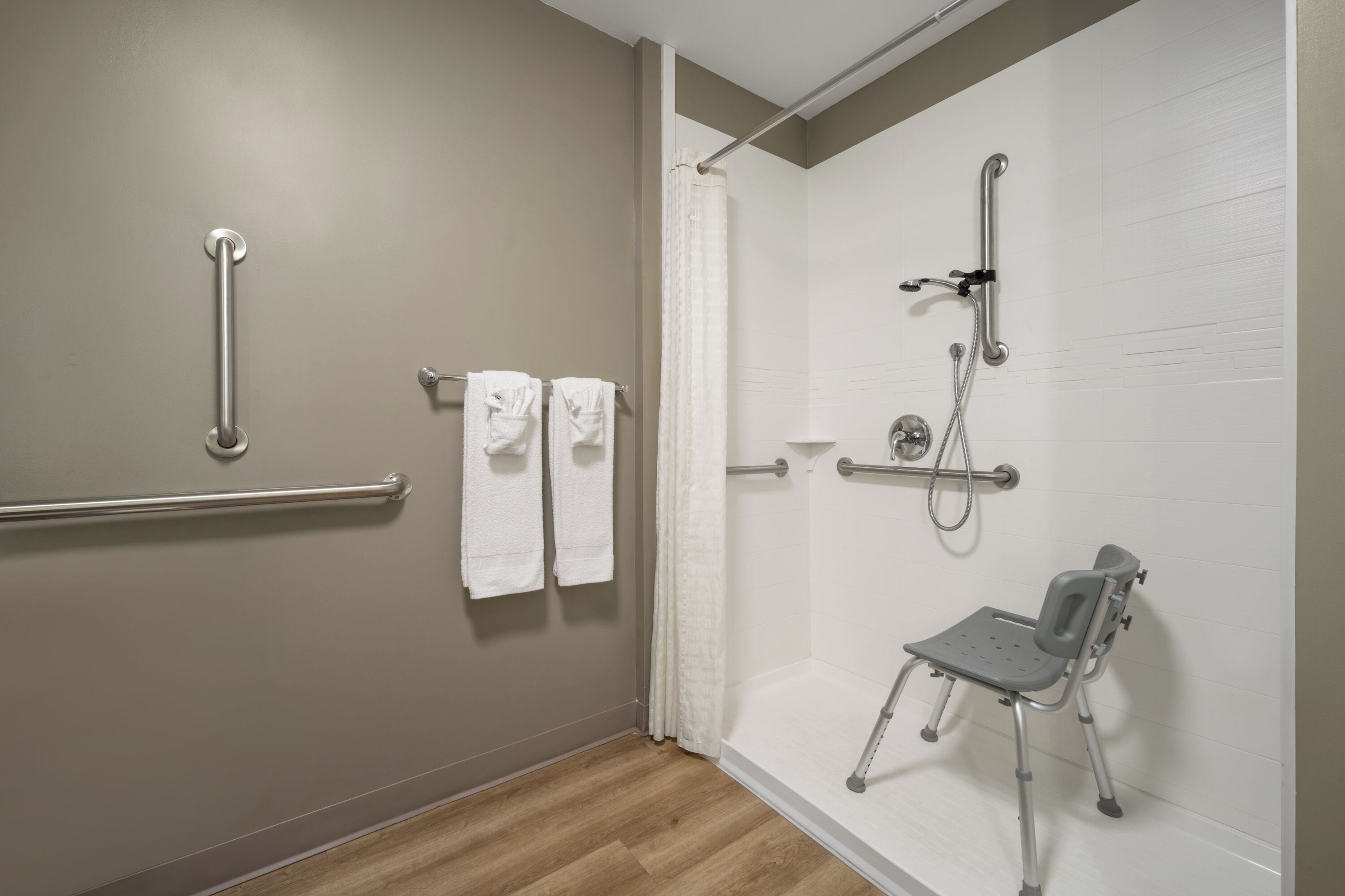 Ein Badezimmer mit Dusche, Haltegriffen, einer rollstuhlgerechten Sitzbank und Handtüchern an einem Haken.