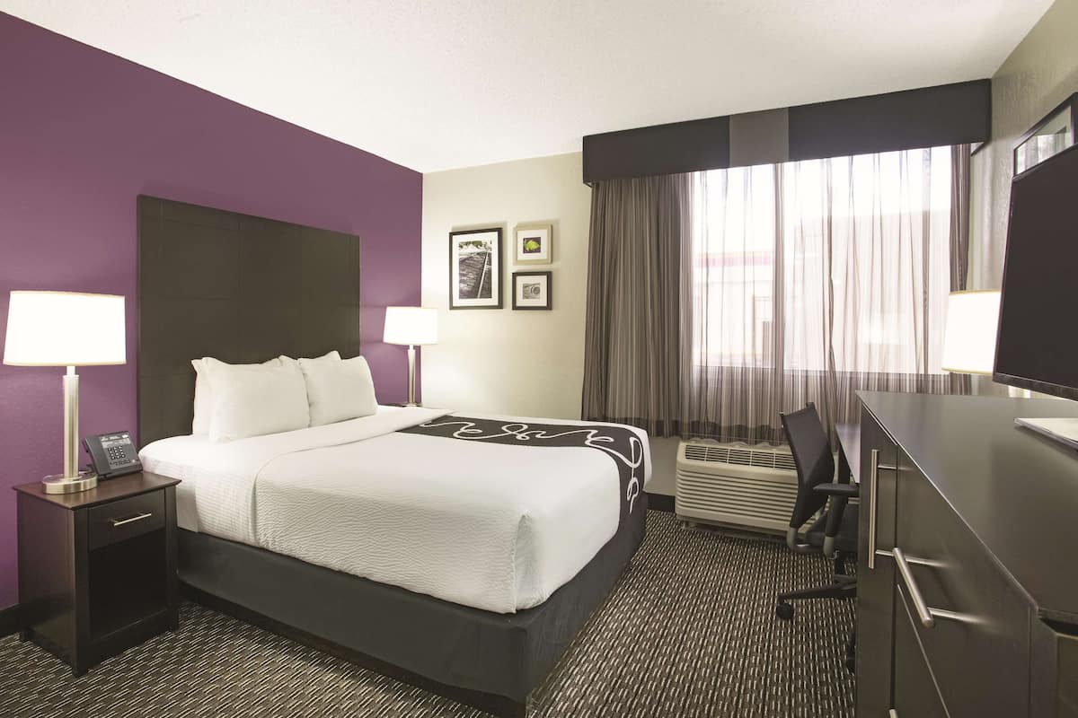 Une chambre d’hôtel avec un grand lit, un bureau et une chaise. Il y a deux tables de chevet avec des lampes et un téléphone. Les murs de la chambre sont de couleur violette et il y a une grande fenêtre avec des rideaux.