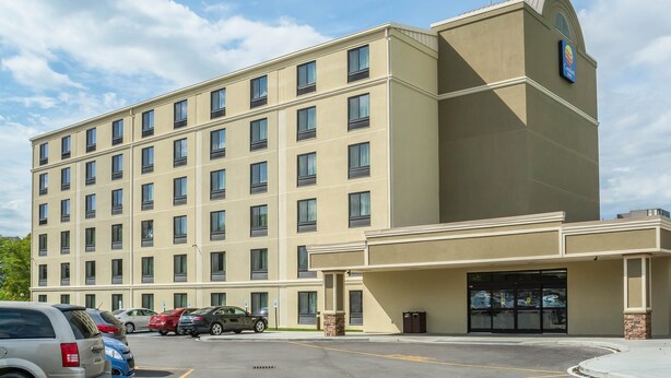 Un hotel de varios pisos con fachada color beige, una entrada amplia con puerta de vidrio y un estacionamiento con varios autos.