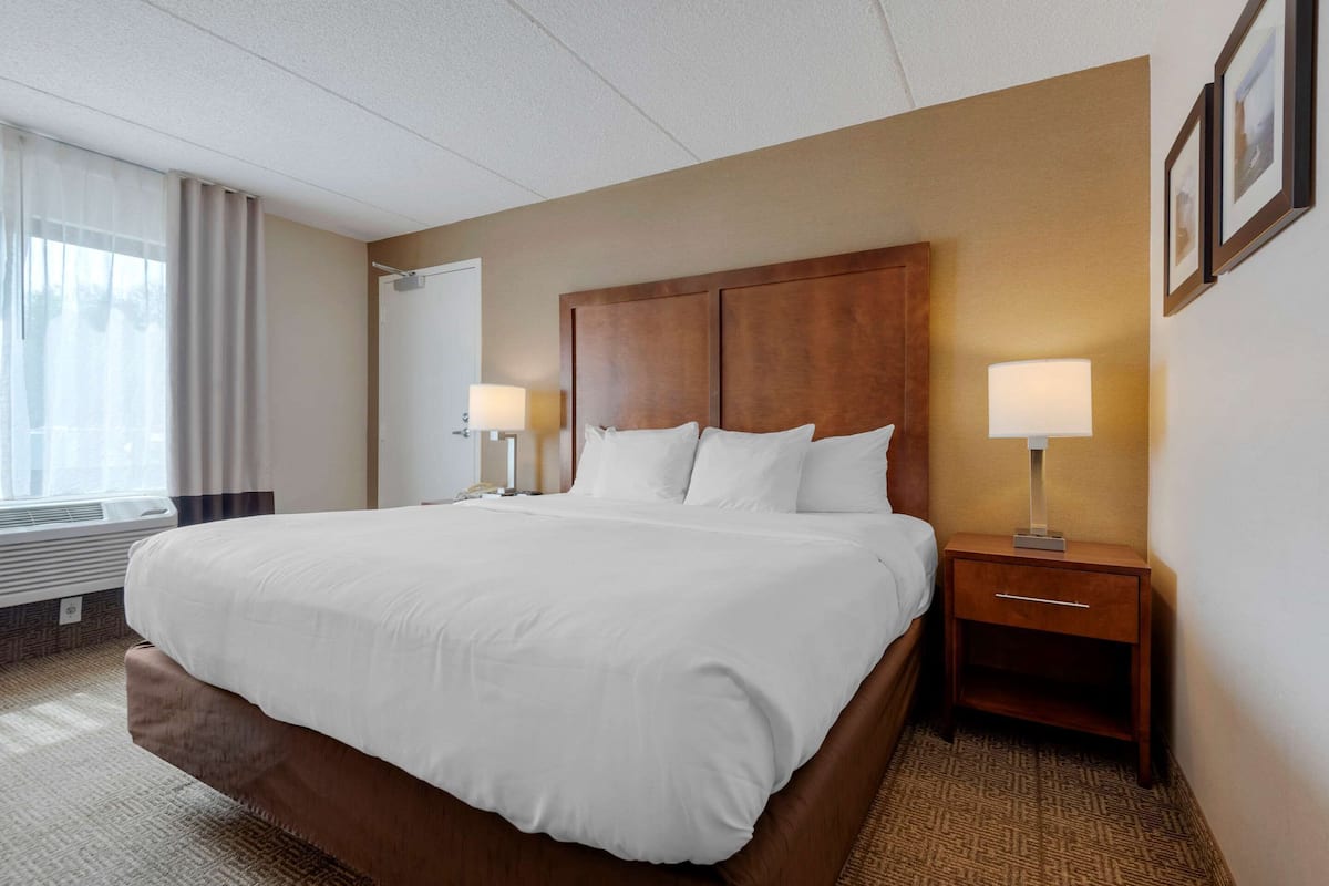 A hotel room with a large bed, two bedside tables with lamps, a window with curtains, and a wall-mounted picture frame.