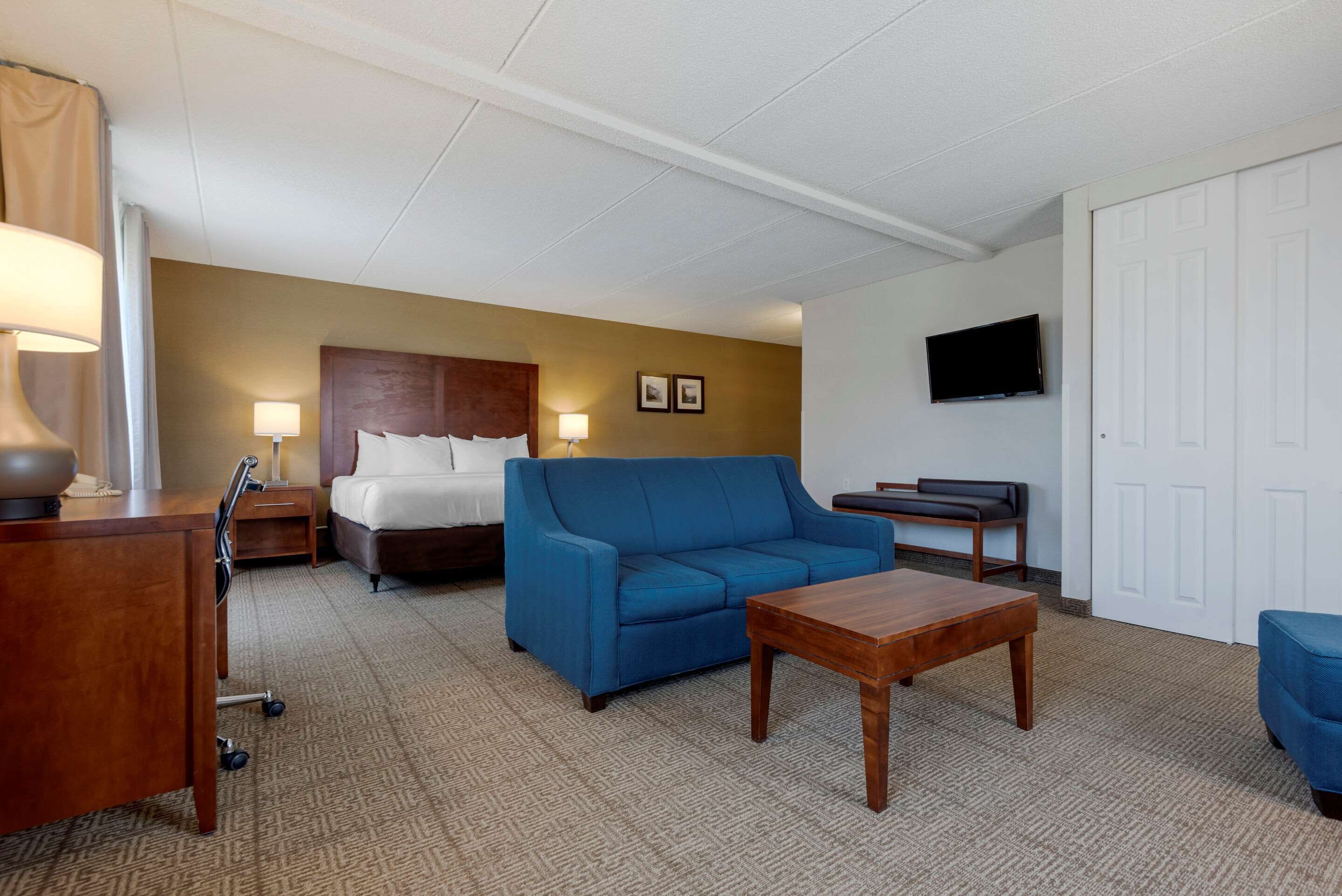A hotel room with a bed, a blue sofa, a wooden coffee table, a desk with a lamp, and a TV mounted on the wall.