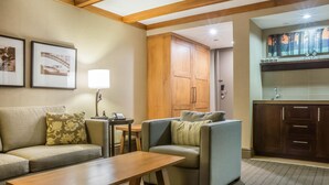 Una sala de estar con un sofá, sillones, una mesa de centro de madera y una lámpara. Incluye una zona de cocina con armarios y un cuadro sobre el fregadero.