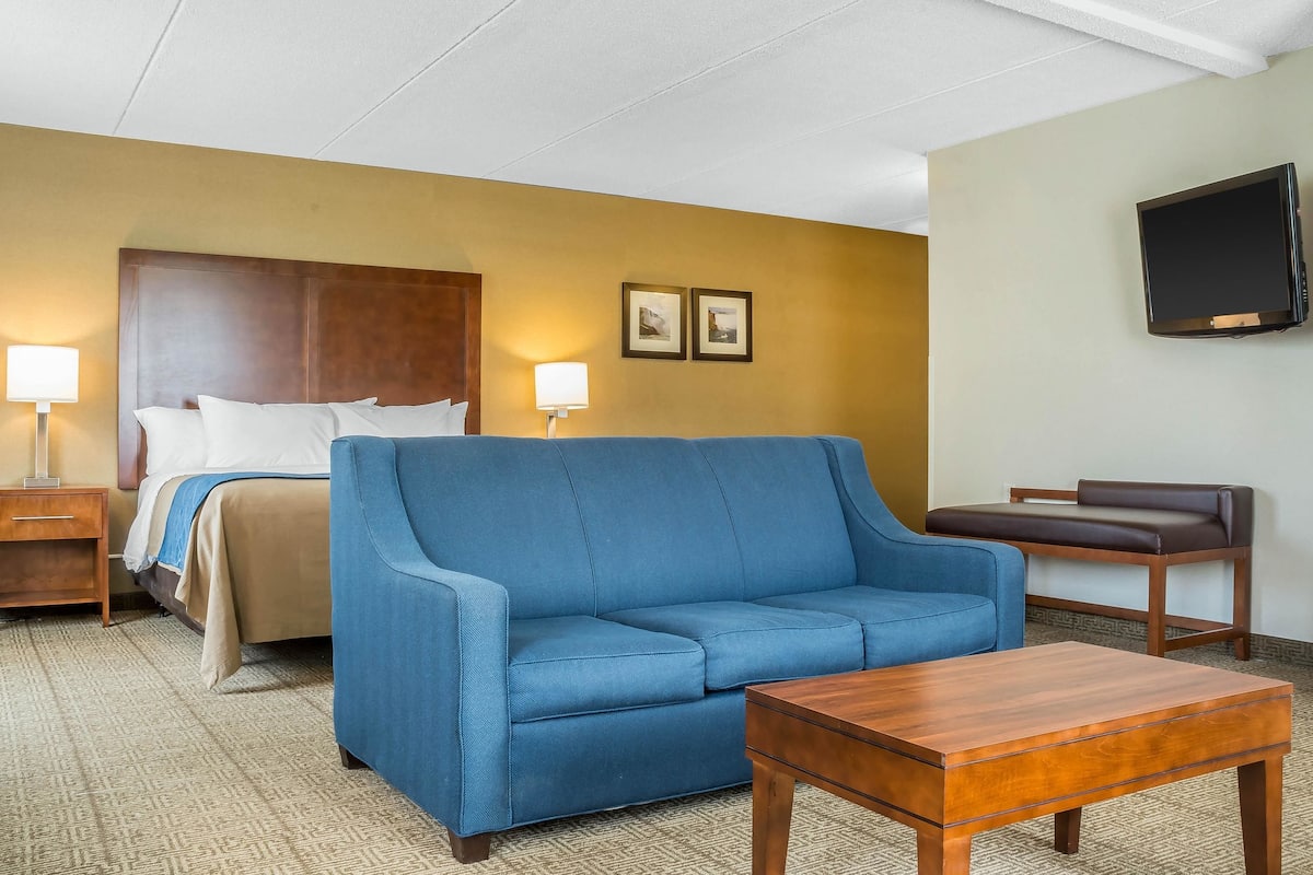 A hotel room with a bed, a blue sofa, a wooden coffee table, a nightstand, a lamp, and a TV mounted on the wall.