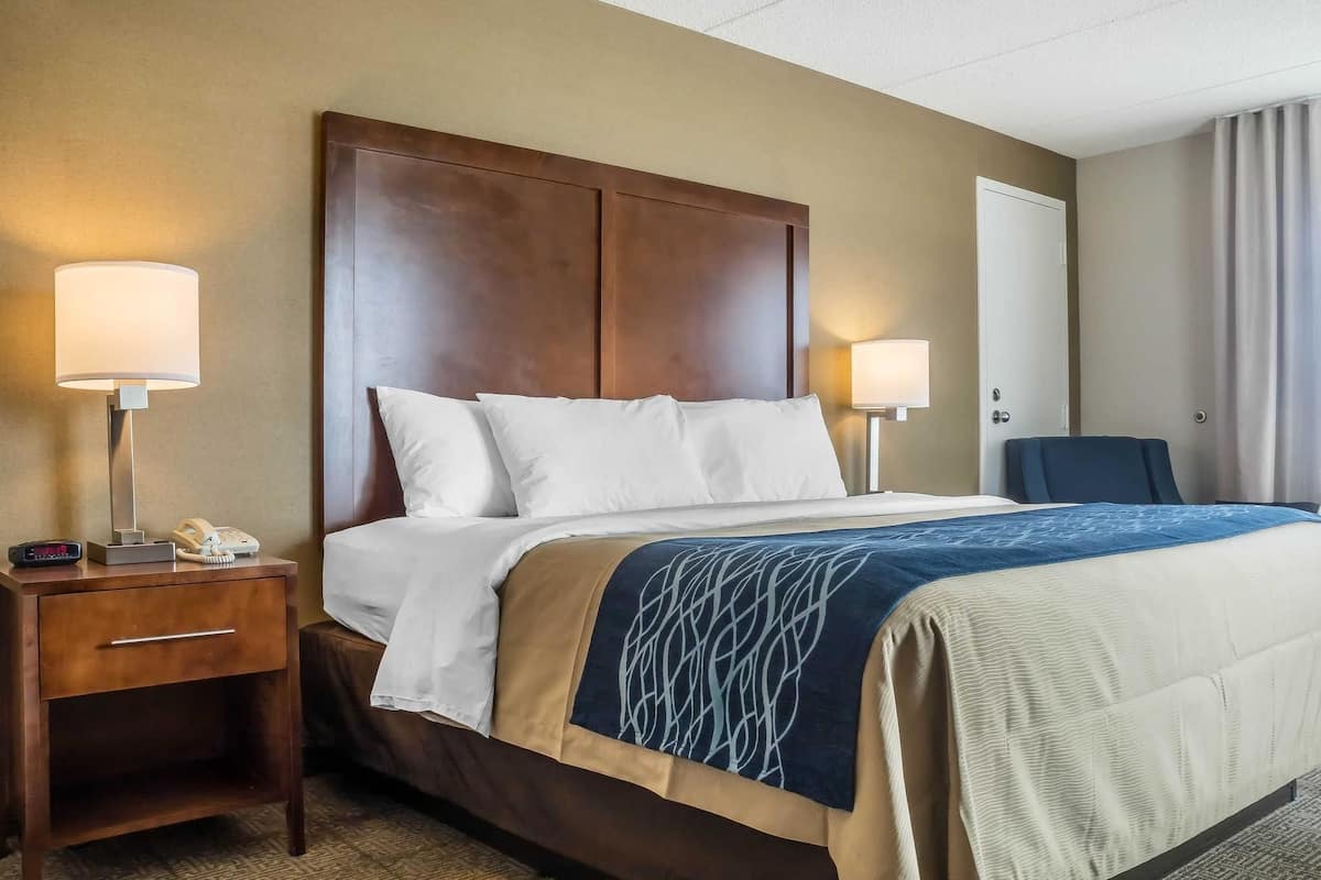 A hotel room with a large bed, two bedside lamps, a nightstand with a phone, and a chair.