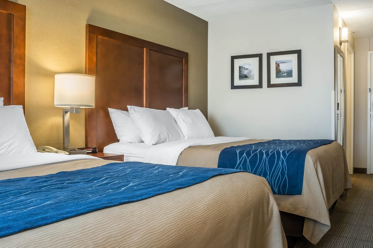 A hotel room with two beds, a wooden headboard, a nightstand with a lamp, and two framed pictures on the wall.