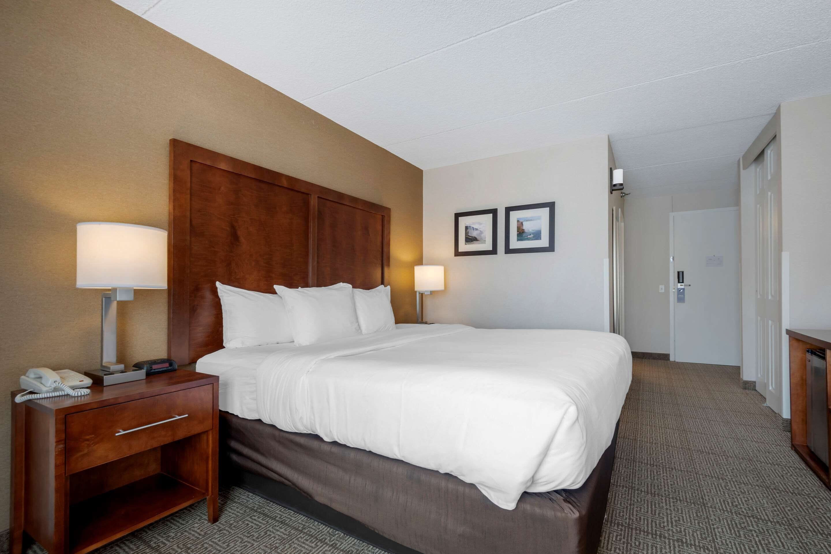 A hotel room with a large bed, a wooden headboard, two bedside lamps, a nightstand with a phone, and a carpeted floor.