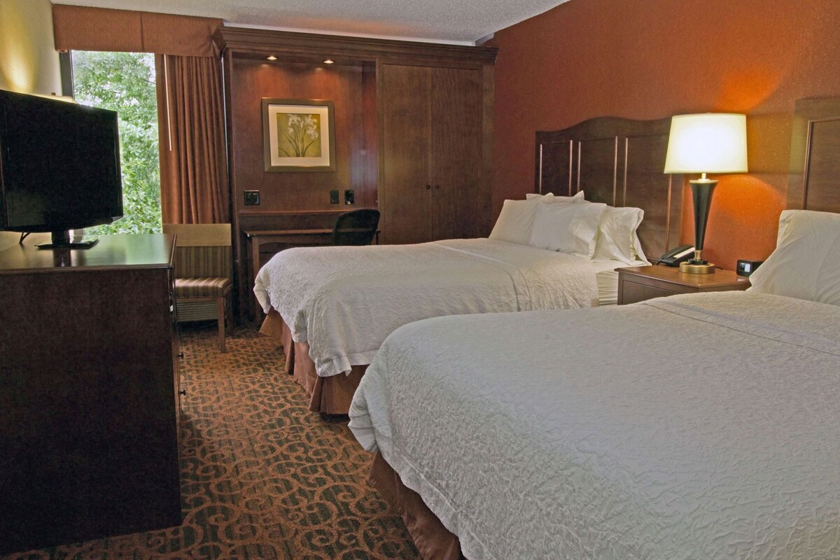 A hotel room with two beds, a desk, a chair, a television, and a lamp.