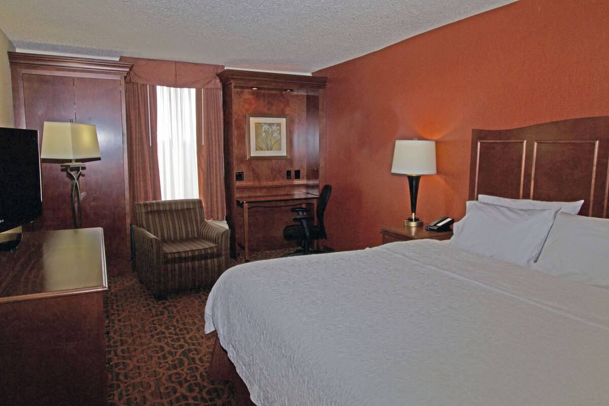 A hotel room with a large bed, a desk, a chair, a television, and a desk lamp.