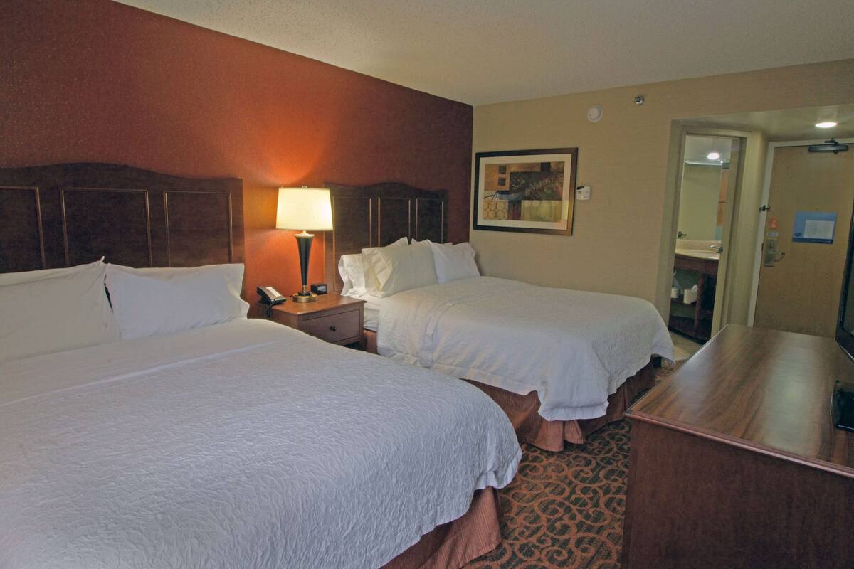 A hotel room with two beds, a nightstand with a lamp, a wooden desk, and a painting on the wall.
