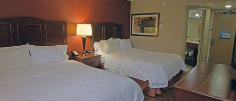 A hotel room with two beds, a nightstand with a lamp, a wooden desk, and a painting on the wall.