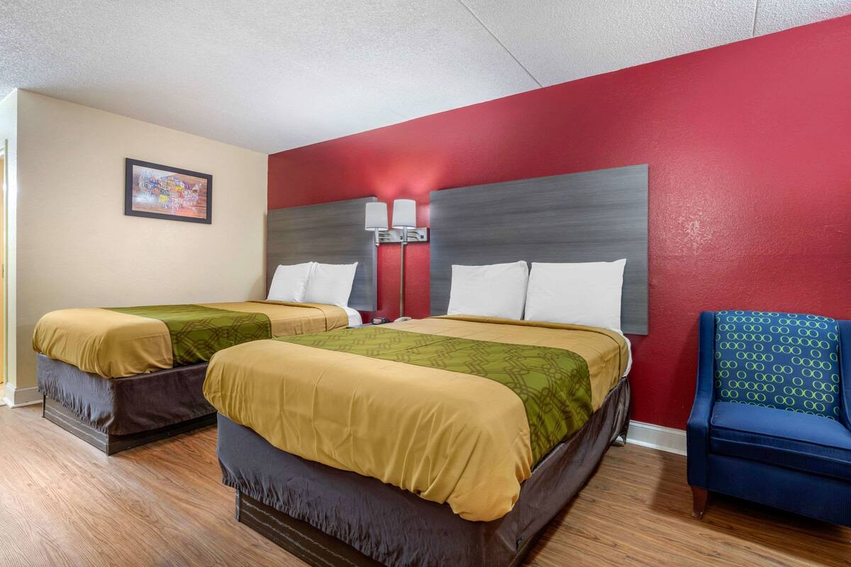 A hotel room with two beds, a red accent wall, a blue chair, and a painting on the wall.