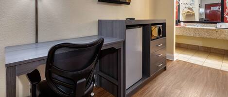 A compact hotel room with a desk, a chair, a television mounted on the wall, a microwave, and a small kitchenette area.