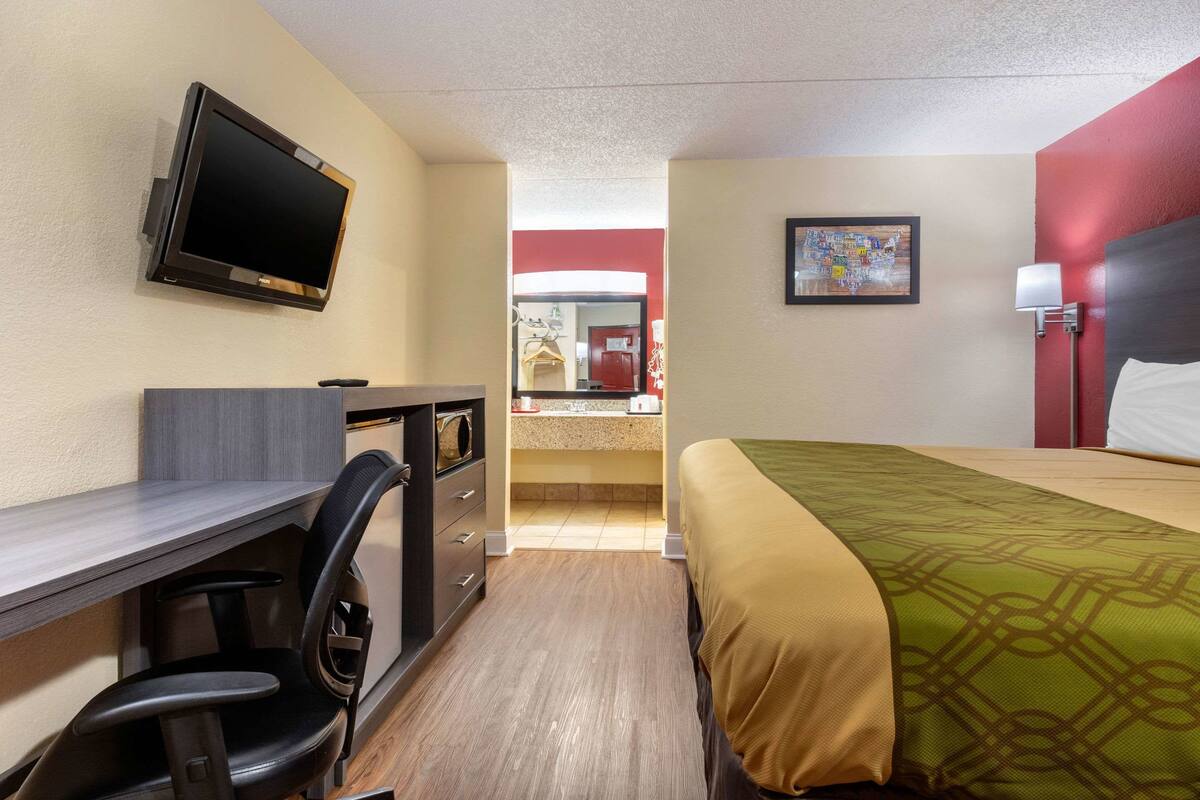A hotel room with a bed, desk, chair, TV, and a small kitchenette.