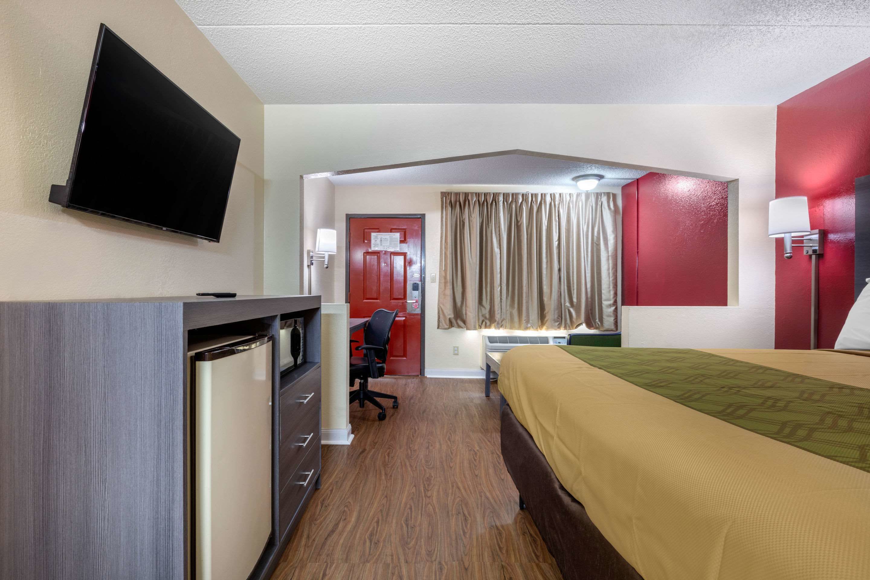 A hotel room with a bed, a desk, a chair, a TV, and a door leading to a bathroom.