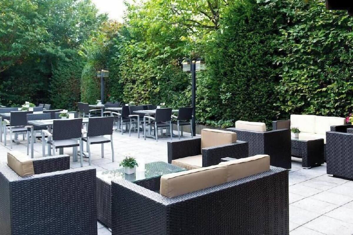 An outdoor patio with wicker furniture, tables, and chairs, surrounded by greenery.