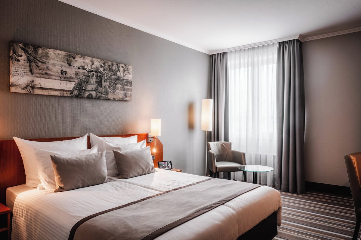 A hotel room with a large bed, a nightstand, a lamp, a chair, and a window with curtains.