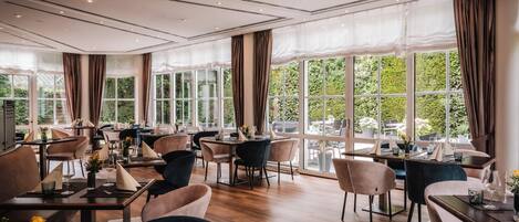 A spacious dining area with multiple tables and chairs, large windows with curtains, and a modern ceiling design.