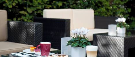 Uma área de estar externa com móveis de vime, uma mesa de vidro e um vaso com planta.