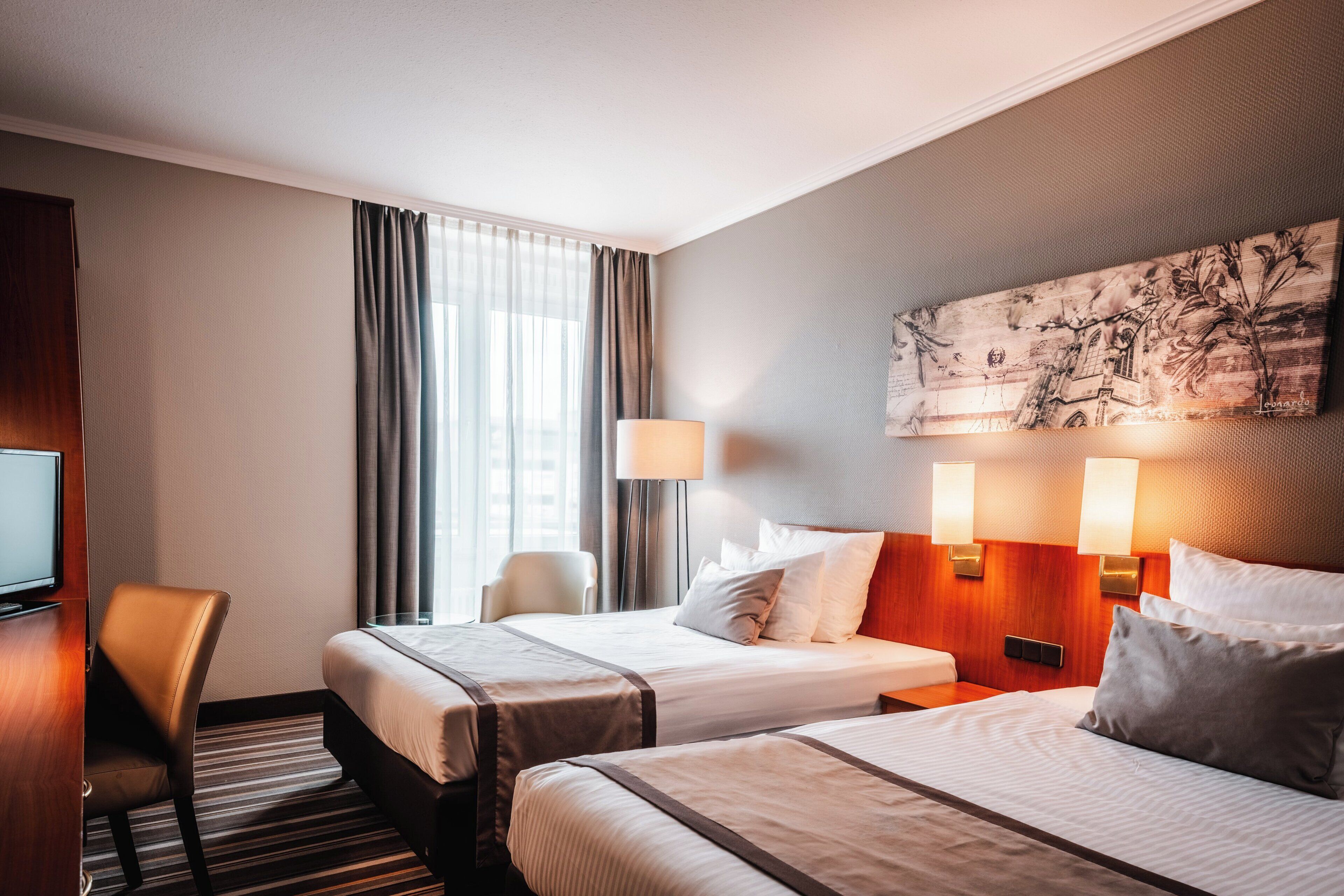 A hotel room with two beds, a desk, a chair, a TV, and a painting on the wall.