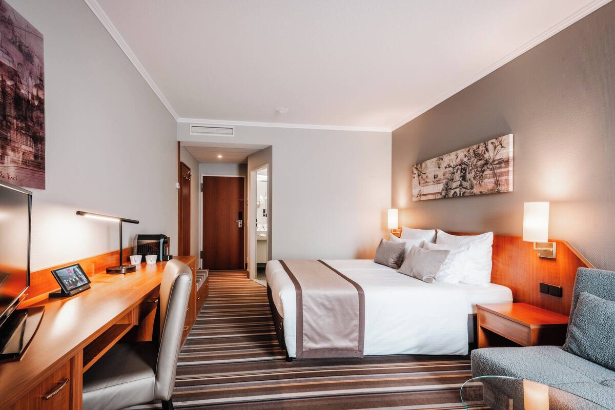 A hotel room with a bed, a desk with a computer, a chair, a bedside table, and a lamp.