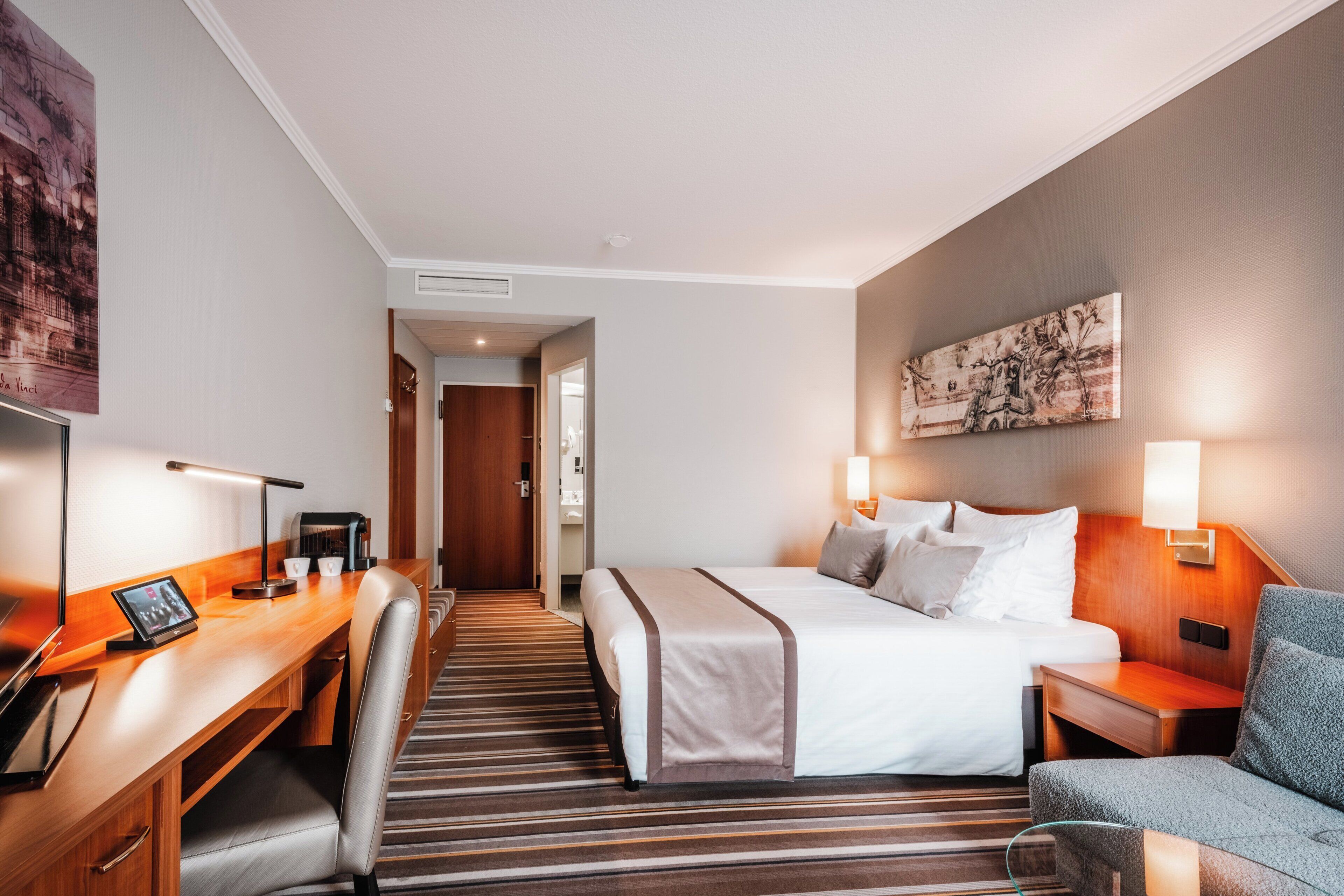 A hotel room with a bed, a desk with a computer, a chair, a bedside table, and a lamp.