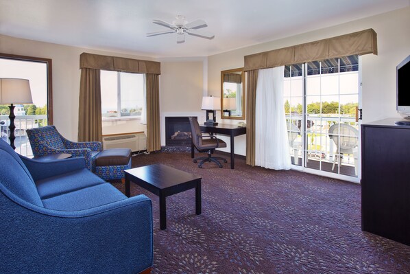 A hotel room with a blue sofa, a small table, a desk with a chair, a television, and large windows with curtains.